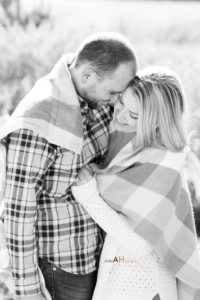 A couple stands close together outdoors at Crissy Fields, wrapped in a plaid blanket, smiling and touching foreheads. The black and white image captures their happy, affectionate moment during an engagement session.