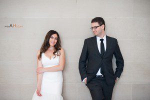A woman in a white dress and a man in a black suit stand against a light-colored stone wall, capturing the joy of their San Francisco elopement. The woman smiles with her arms crossed as the man looks at her with his hands in his pockets.