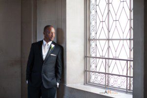 A man in a black suit with a white shirt and boutonniere stands by a large window with decorative glass, looking thoughtfully outside in the softly lit room of a San Francisco City Hall Wedding.