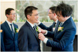 Four men in suits, smiling. One man pins a boutonniere on another’s jacket while two others watch, all in a bright room. It appears to be San Ramon Waters wedding preparation or another formal event.