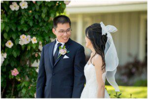 A bride and groom stand outdoors at a San Ramon Waters wedding, smiling at each other. The groom wears a navy suit with a boutonniere, and the bride wears a white dress with a bow in her hair. Flowering greenery fills the background.