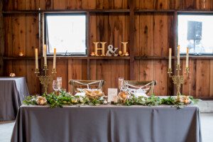 A rustic wedding sweetheart table with a gray tablecloth, decorated with greenery, candles, and gold candelabras. Behind the table are wooden walls and gold initials H & J on the window ledge.
