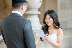 A woman in a white dress smiles and reads from a piece of paper during a wedding ceremony, standing opposite a man in a suit inside an elegant, ornate building.