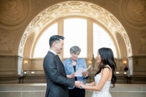 A couple stands facing each other, holding hands and smiling, while an officiant reads from a booklet in a grand, ornate hall with arched windows behind them.