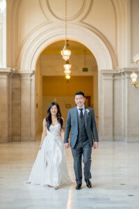A bride in a white dress and a groom in a gray suit walk hand in hand, smiling, in a grand hallway with marble floors and arched ceilings with hanging lights.