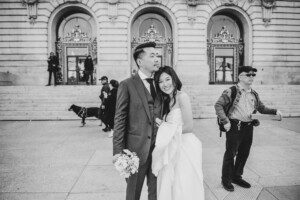 A bride and groom stand smiling together outside a historic building with ornate doors. The bride holds the grooms arm, holding a bouquet. Several people and a dog are in the background. The image is in black and white.