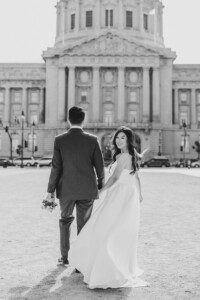 A bride in a flowing white dress smiles while holding hands with the groom, who is holding a bouquet. They stand outside a grand building with columns, shot in black and white.