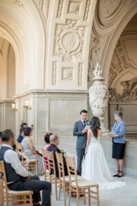 A bride and groom stand facing each other during their wedding ceremony in an ornate hall with arched ceilings. Guests are seated nearby, and an officiant stands beside the couple, holding a book.