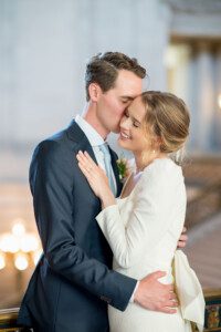 A bride and groom embrace and smile as the groom kisses the bride on the forehead. The bride wears a white dress, and the groom wears a dark suit and light blue tie. They stand in a softly lit, elegant indoor setting.