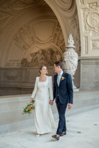A bride in a long-sleeved white dress and a groom in a navy suit walk hand in hand, smiling at each other, inside an ornate building with detailed arches and marble floors. The bride holds a bouquet of flowers.
