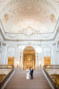 A bride and groom stand together on grand marble stairs beneath an ornate, domed ceiling in an elegant, historic building. The bride holds a bouquet, and the couple gazes at each other, surrounded by intricate architectural details.