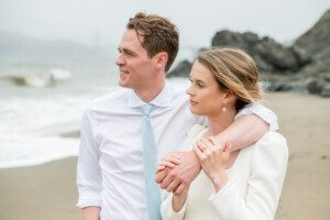A couple dressed in white stands close together on a beach, with the man’s arm around the woman’s shoulders. They both look into the distance, the sea and rocky shore visible behind them.