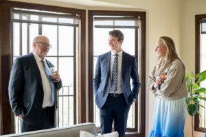 Three people dressed in formal attire stand indoors near large windows, smiling and holding drinks, appearing engaged in a cheerful conversation. A green potted plant is visible beside them.