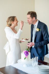 A bride in a white dress feeds cake to a groom in a navy suit with a boutonnière. They stand by a table with a white cake topped with pink flowers and two empty champagne flutes, both smiling and enjoying the moment.