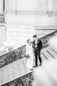 A bride and groom, dressed elegantly in wedding attire, stand together on a grand staircase, looking at each other and smiling. The setting features ornate railings and classical architecture.