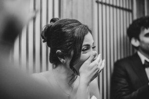 A woman in formal attire, smiling and covering her mouth with her hand, appears joyful. She is indoors, with a blurred person in a suit beside her and vertical wooden panels in the background. The image is in black and white.