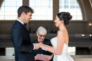 A bride and groom face each other, smiling and holding hands during a wedding ceremony, while an officiant stands behind them holding a book. Sunlight streams through large arched windows in the background.