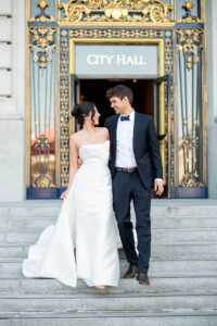A bride in a white dress and a groom in a black suit smile at each other while standing on the steps outside a building with ornate gold doors and a sign that reads City Hall.