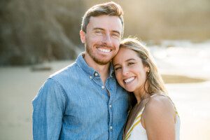 A smiling couple stands close together on a beach, with the woman resting her head on the mans shoulder. The sun shines brightly in the background, creating a warm and happy atmosphere.
