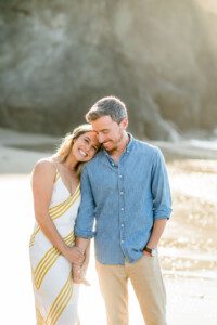 A smiling woman in a white dress with yellow stripes leans her head on a mans shoulder. The man, in a blue button-up shirt, gently holds her hand. They stand together on a sunlit beach with rocks in the background.