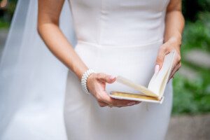 A bride in a white dress and pearl bracelet holds and reads a small open book outdoors, with greenery blurred in the background.