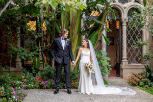 A bride and groom stand hand in hand outdoors, dressed in a white wedding gown and black tuxedo, surrounded by lush green plants and flowers, in front of an ornate stone building with arched windows.