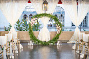 An elegant wedding ceremony setup features a large circular greenery and pink flower arch, white draped curtains, gold chairs, and white candles arranged along the aisle on a polished floor.