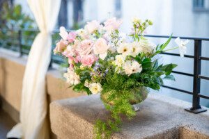 A silver vase filled with pink and white flowers and green foliage sits on a stone balcony ledge beside a sheer white curtain, with a railing and blurred background in view.