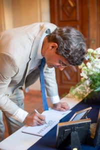 A man in a beige suit and blue tie leans over a table, signing a book with a pen. A framed photo and a floral arrangement are on the table beside him. The setting appears formal and indoors.