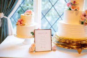 Two elegant white cakes decorated with pink flowers are displayed on stands near a window. A dessert bar menu with floral accents sits between them on the table.
