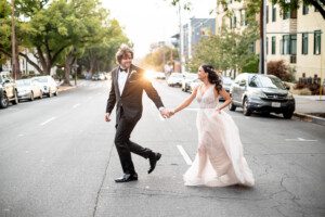 A bride and groom hold hands and smile while walking across an empty city street, surrounded by parked cars and trees, with sunlight shining between them.