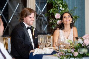 A man in a black tuxedo and a woman in a sleeveless dress sit together at a decorated table, both smiling and laughing, surrounded by flowers and glassware at what appears to be a formal event or celebration.