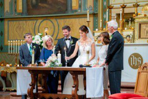 A bride and groom stand at a table in a church, lighting a unity candle together, surrounded by four older adults, likely family members, during a wedding ceremony. Everyone is dressed formally.