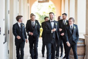A group of men in black suits and bow ties, some laughing and smiling, walk down a bright, elegant hallway with arches and wooden barrels.