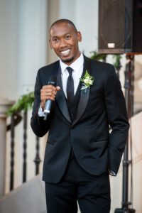 A man in a black tuxedo with a boutonnière smiles while holding a microphone, standing indoors near a staircase decorated with greenery.