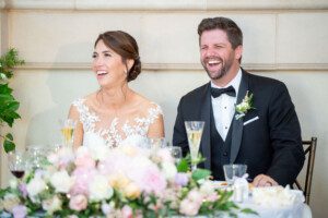 A bride in a white lace dress and a groom in a black tuxedo sit side by side, laughing joyfully at a decorated table with flowers and champagne glasses in front of them.