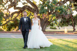 A bride in a white gown and a groom in a black tuxedo walk hand in hand on grass, smiling, with a large leafy tree in the background on a sunny day.