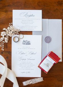 Flat lay of elegant wedding invitations, RSVP card, silver rings, a jeweled hairpiece, a wax seal on an envelope, a lace bracelet, white ribbon, and an engagement ring in a red velvet box on a wooden table.