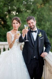 A bride in a white lace wedding dress and a groom in a black tuxedo stand outdoors, smiling and raising champagne glasses in a toast. Lush green trees are in the background.