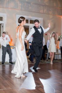 A bride and groom dance joyfully on a wooden floor at their wedding reception, surrounded by cheering guests. The bride wears a white gown and the groom wears a black vest and pants.