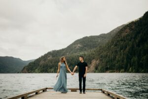 A couple holds hands while standing on a wooden dock over a lake, surrounded by forested mountains under a cloudy sky. The woman wears a flowing blue dress and the man wears a black shirt and pants.