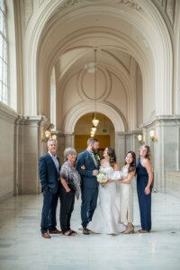 A bride and groom stand together in an elegant hall with high arched ceilings, surrounded by four smiling guests posing for a group photo.
