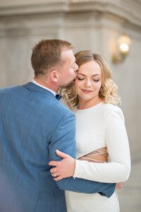 A man in a blue suit kisses a woman in a white dress on the temple. The woman smiles softly with her eyes closed. They embrace in a softly lit, elegant indoor setting.