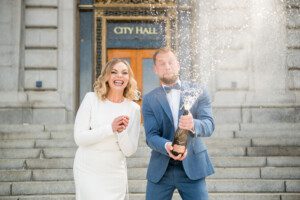A joyful couple, dressed in formal attire, stands on city hall steps. The man pops a champagne bottle with a burst of spray, while the woman smiles brightly beside him.