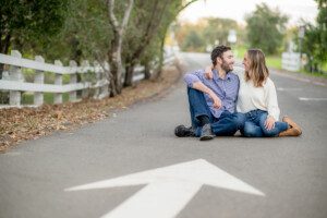 A couple sits together on a quiet road, smiling at each other. They are surrounded by trees and a white fence, with a large white arrow painted on the pavement pointing toward them.