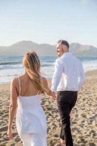A couple walks hand in hand along a sandy beach, both dressed in white. The woman has long blonde hair, and the man looks back smiling. Ocean waves and mountains are visible in the background under a clear sky.
