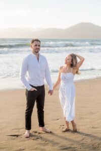 A man and woman stand barefoot on a sandy beach near the ocean, holding hands. The man wears a white shirt and black pants; the woman wears a white dress and smiles while touching her hair. Mountains and waves are in the background.