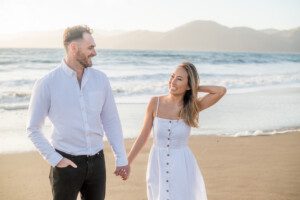 A man and woman holding hands and smiling at each other while walking on a sandy beach, with gentle ocean waves and mountains in the background. Both are wearing white outfits.