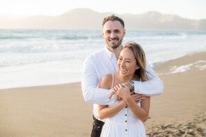 A smiling couple stands on a sandy beach with mountains and waves in the background. The man, in a white shirt, hugs the woman, who is also in white, from behind as she laughs. Both look happy and relaxed.