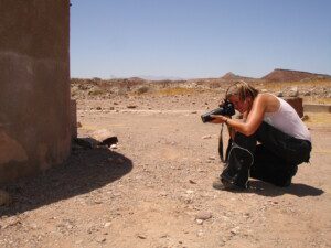 A person wearing a headscarf, white tank top, and dark pants crouches on rocky ground in a desert landscape, holding a camera and photographing a building wall under a clear blue sky.